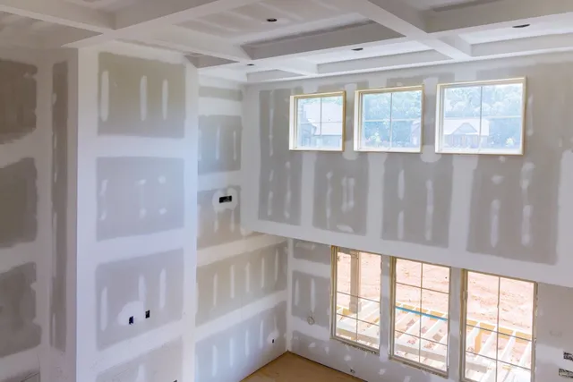 Unfinished drywall interior with taped seams and large windows letting in daylight.