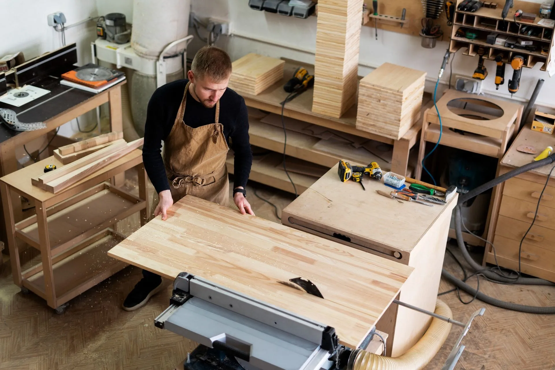 Person in an apron measuring wood in a cluttered workshop with tools and lumber.