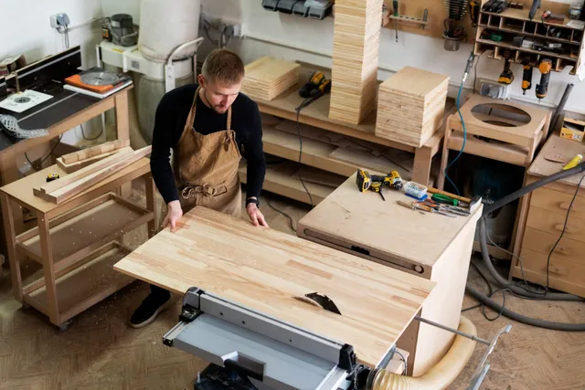 Person in an apron measuring wood in a cluttered workshop with tools and lumber.
