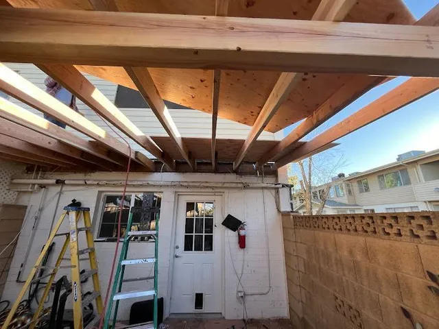 Backyard patio under construction with exposed wooden roof beams, ladder, and tools against a stucco wall
