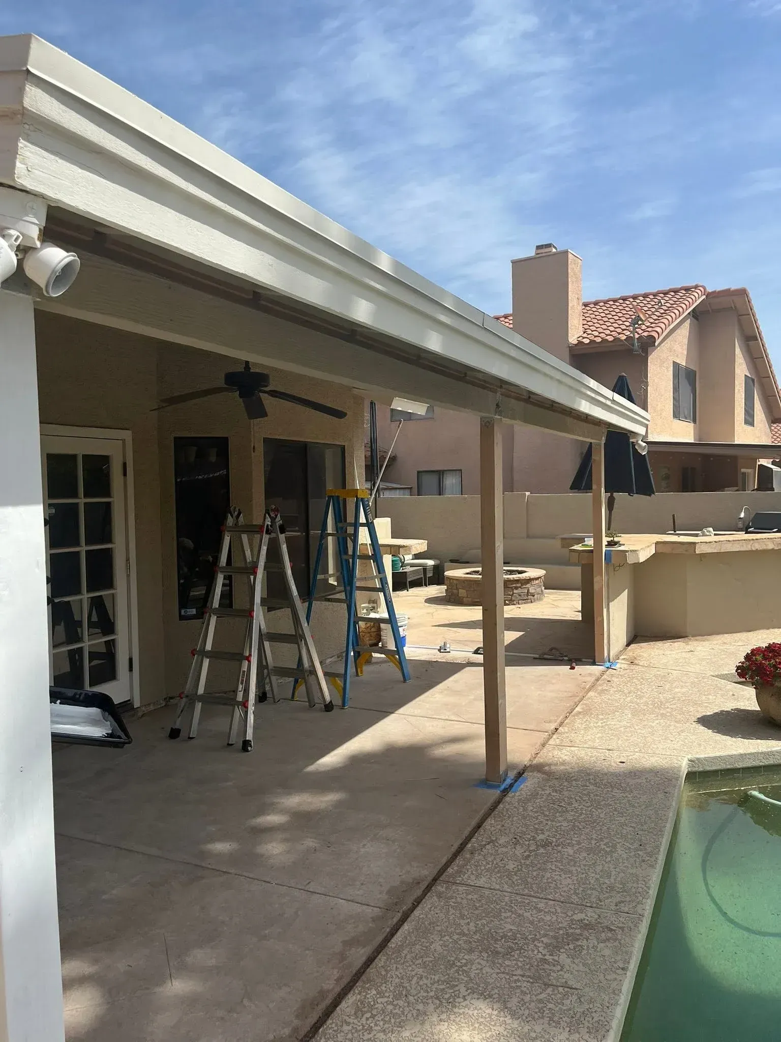 Covered patio with ladders and tools beside a backyard pool under construction