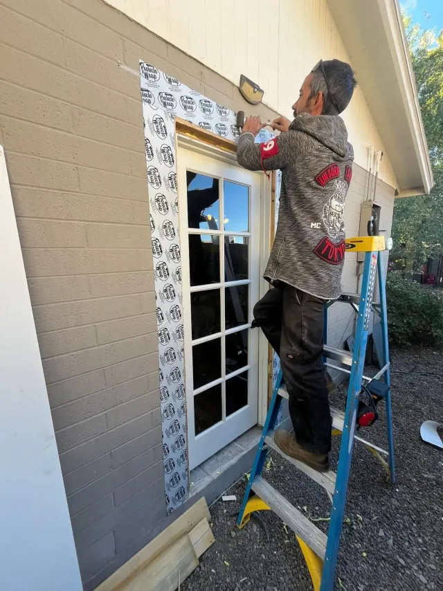 Person on a ladder installing trim around a window on a beige house exterior.