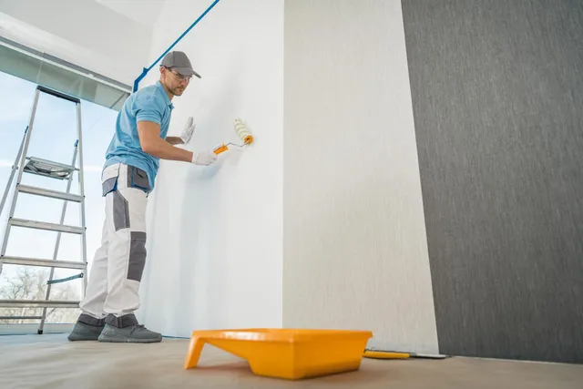 Painter rolling white paint on a wall in a bright room, with a ladder and paint tray nearby