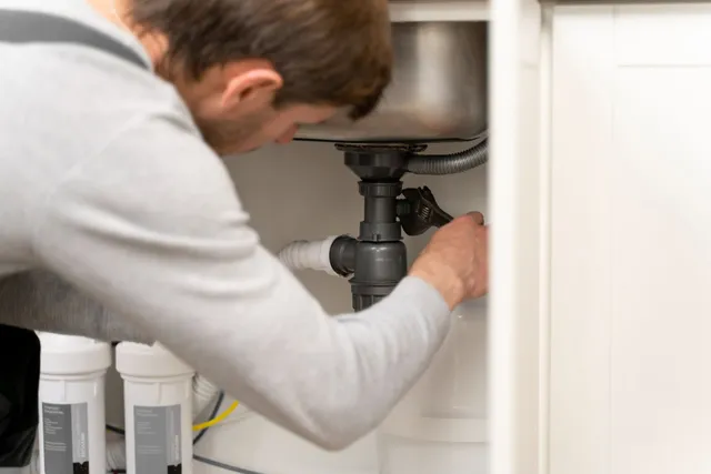 Person fixing plumbing under a sink with tools and cleaning bottles nearby
