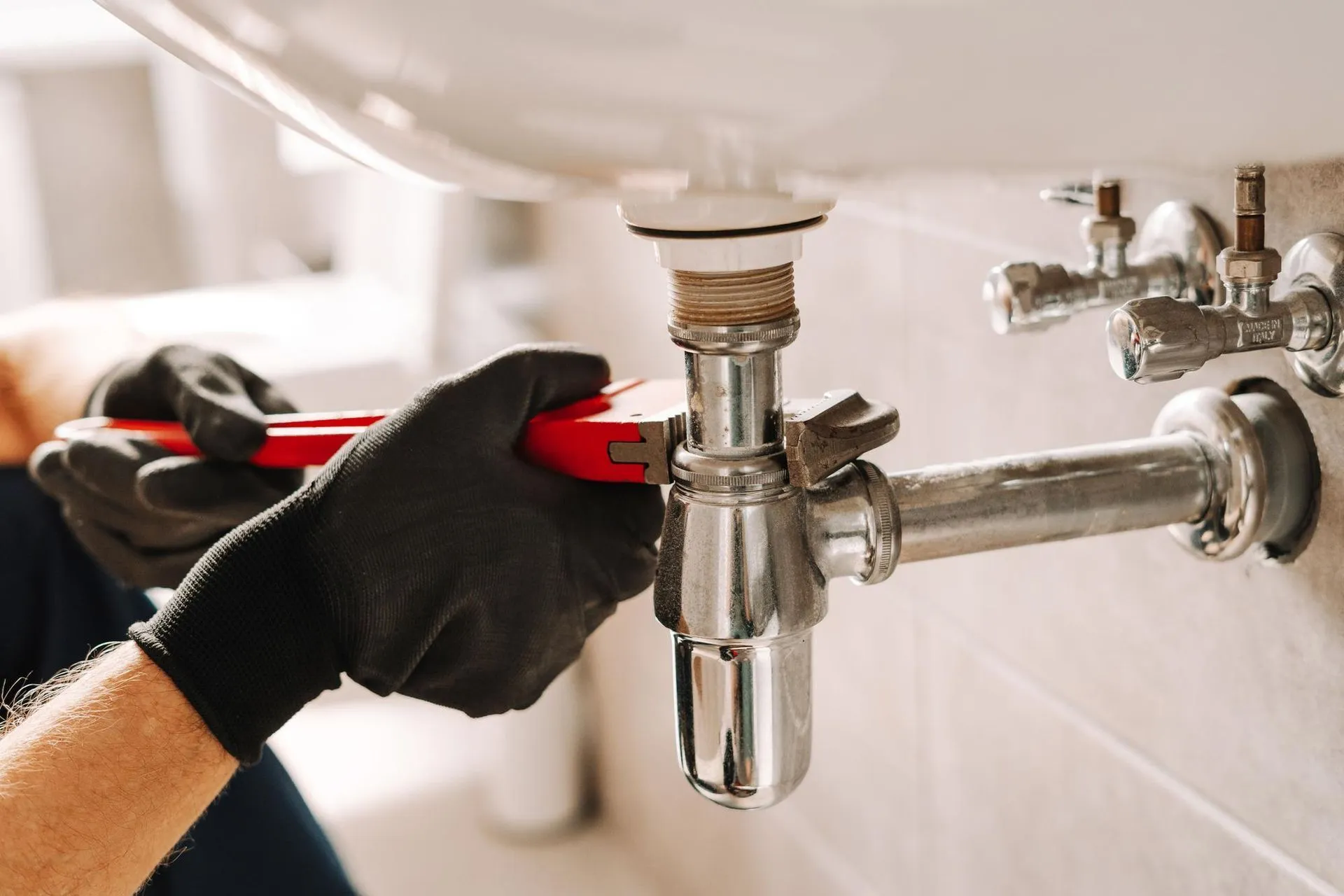 Gloved hands using a wrench to tighten a chrome sink drain pipe under a bathroom sink